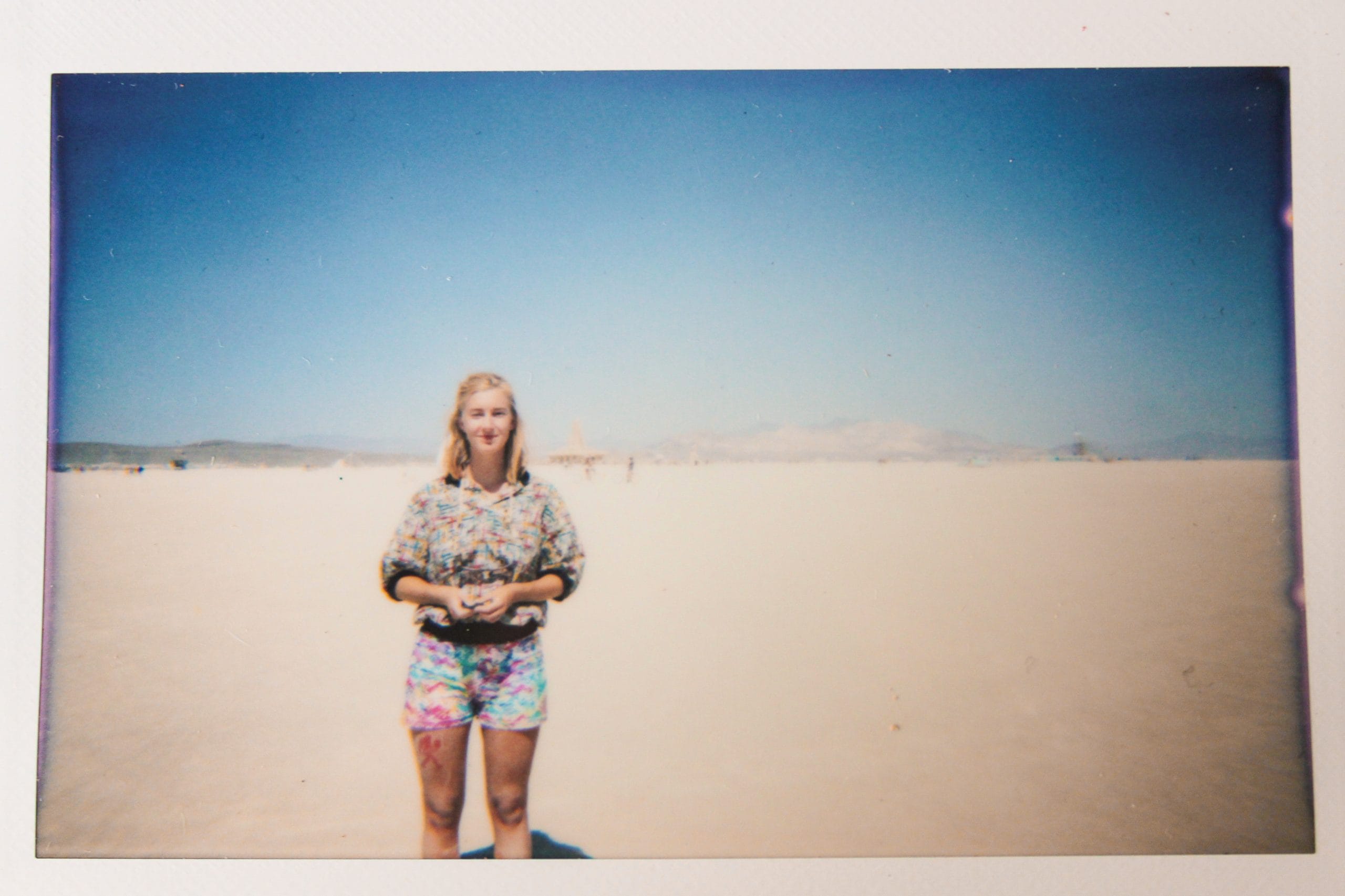 Me, Tasha, looking into the camera lens and standing alone at Burning Man with only the dusty ground in the distance. Tasha is wearing vibrant and patterned clothing and very tan and dusty.