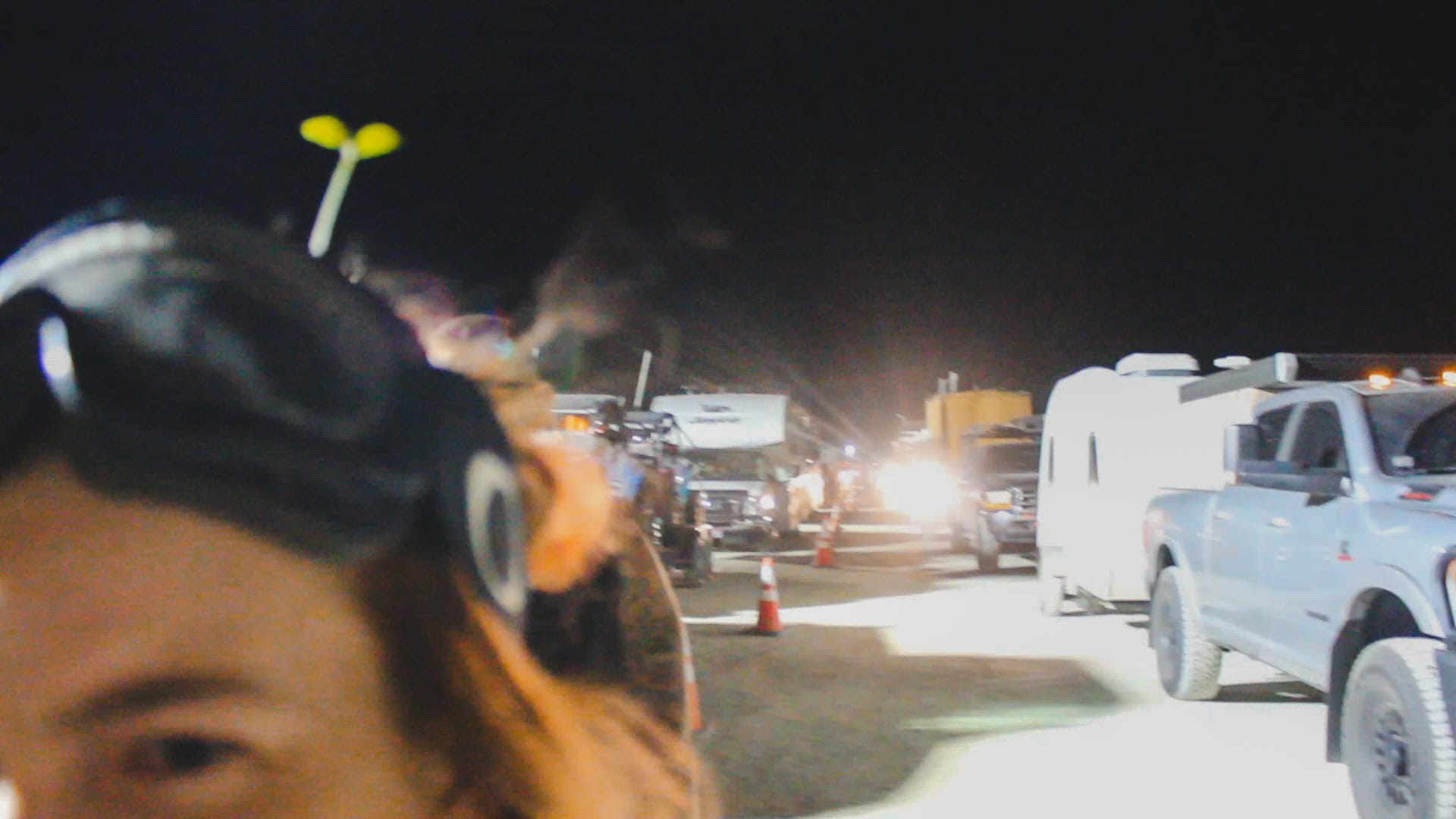 Tasha on the left looking ahead with dust goggles on her head and a small sprout hair clip. Behind her is two lines of cars divided by traffic cones at night time, waiting to get into Black Rock City at the entrance Gate during a volunteer experience.