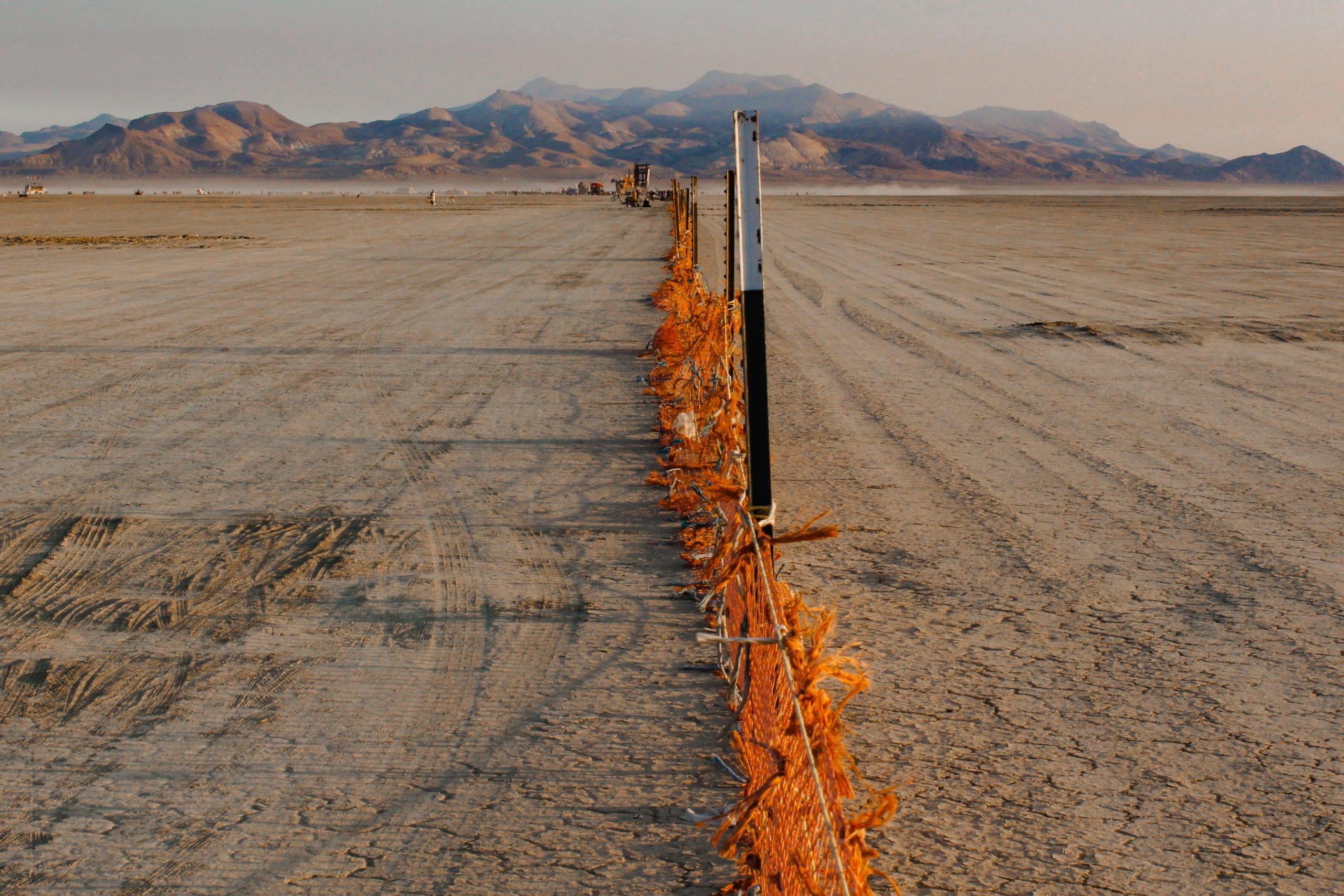 The orange netted trash fence at Burning Man leading a straight line to the mountain hills in the distance. Sunrise time so the dust on the ground and fence are warmly illuminated.