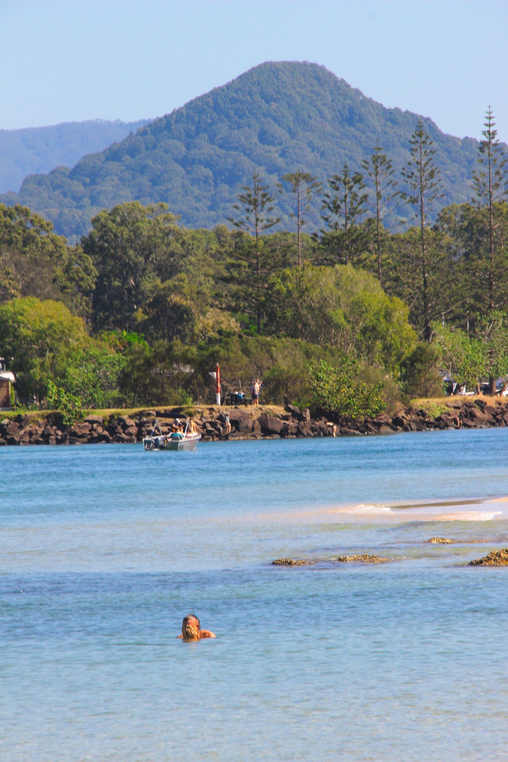 A view of Mount Chincogan from Christmas Beach. Light blue turquoise water then same trees in the distance and above is the mountain greenery.