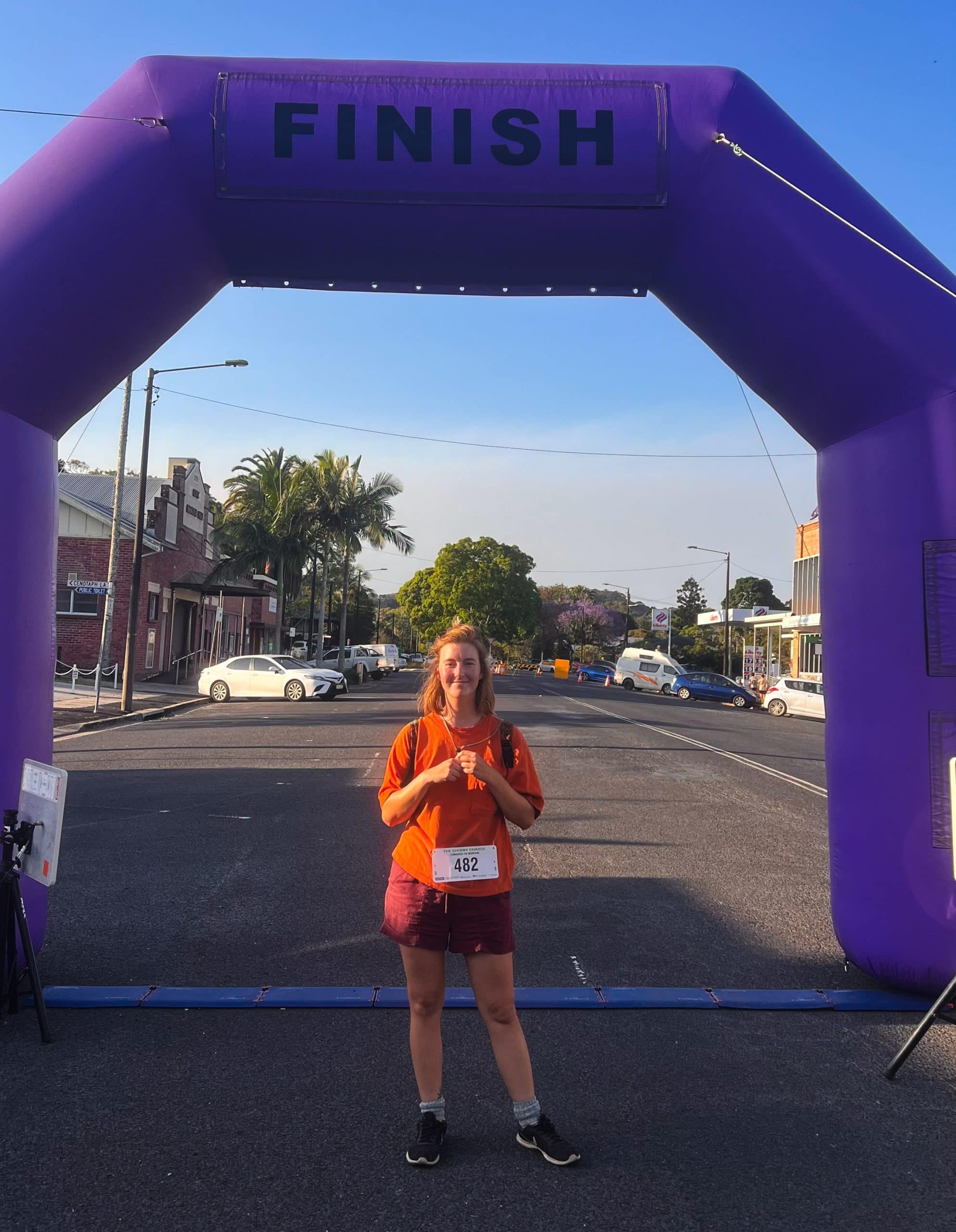 Me looking into the camera wearing bright orange and looking sweaty and flushed. She is alone under the purple inflatable arch finish line for the Chinny Charge hike.