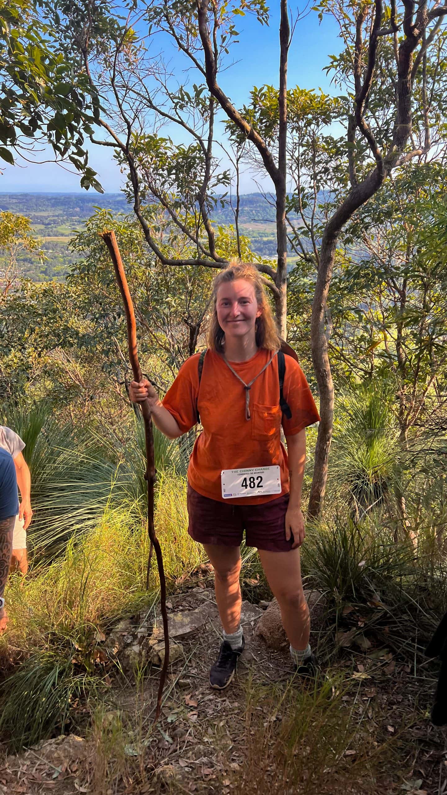 Tasha at the top of Mount Chincogan with trees behind her then a view of the valley below. She is looking sweaty, flushed, and holding a walking stick looking into the camera.